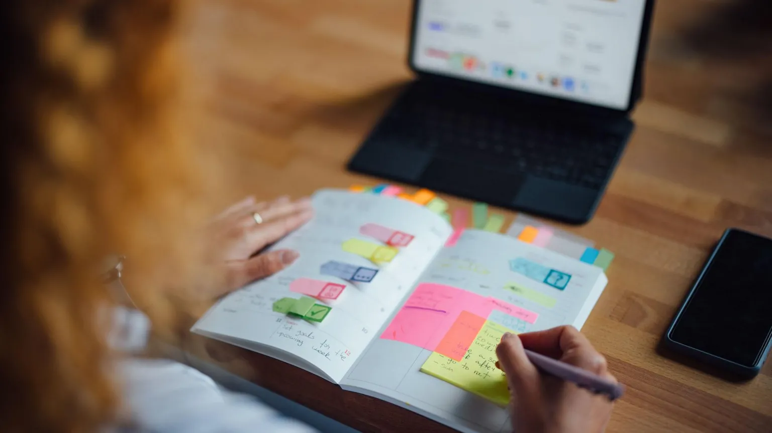  A person with their diary in their hand while looking at a tablet propped up on a table. The diary has lots of colourful tabs and post-its in it