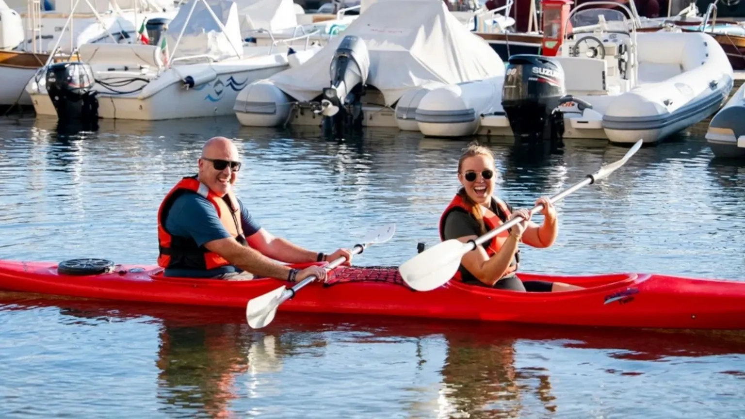 BBC/Studio Lambert a man and woman on a tandem kayak, holding their paddles out of the water and smiling at the camera