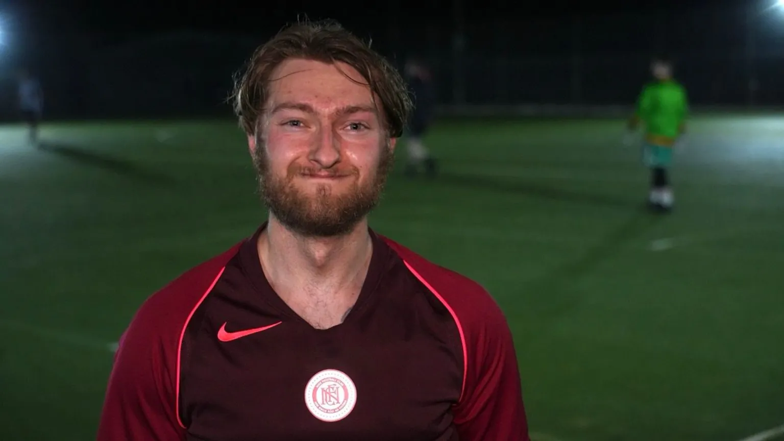 John Fairhall/BBC Lewis has dark, sweat-soaked hair and a ginger beard. He is wearing a red football shirt and standing on a green football pitch. 