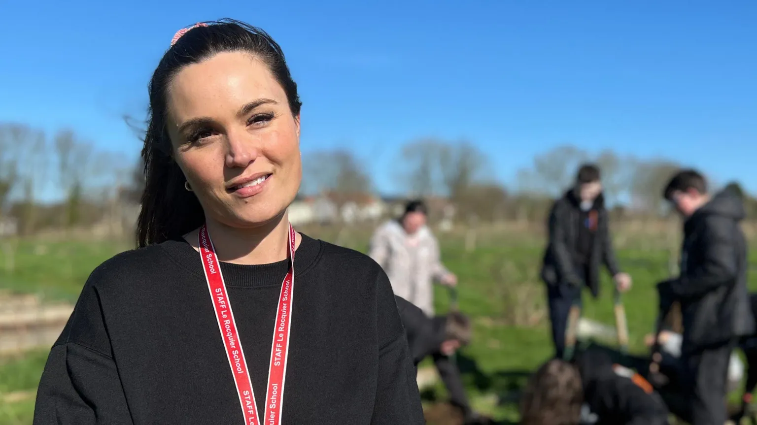 Ellen is wearing a black sweater and red lanyard stands in front of a group working in a garden, with others digging and preparing soil in raised beds in the background.