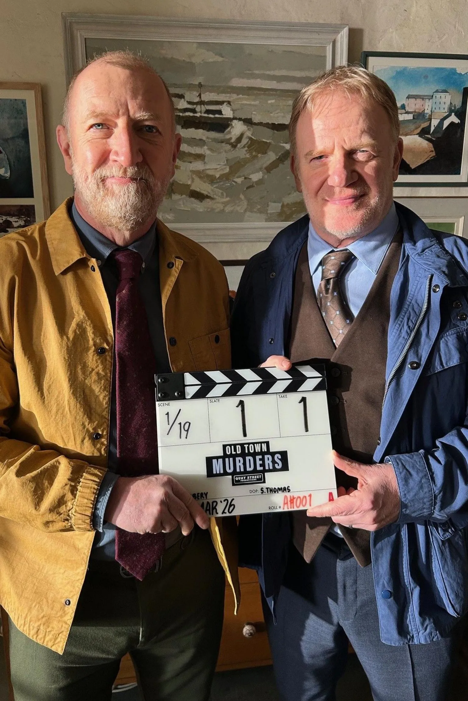 Quay Street Productions Steffan Rhodri and Mark Lewis Jones holding a clapperboard between them. Rhodri is on the left in a mustard jacket and a burgundy tie, with a blue shirt, while Jones is on the right wearing a blue jacket, blue shirt and brown waistcoat and tie. They are both stood up. 