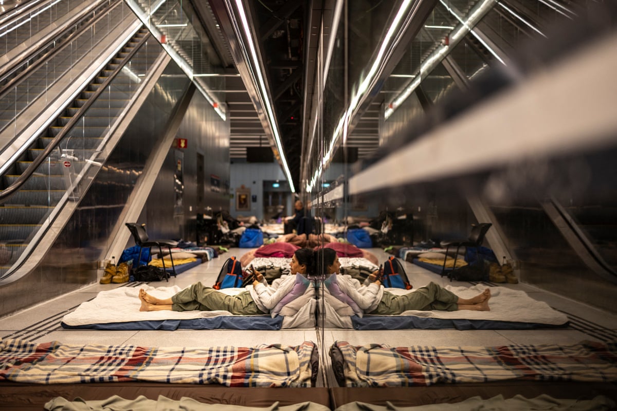 People lying on the blanketed floor of a railway station next to an escalator.
