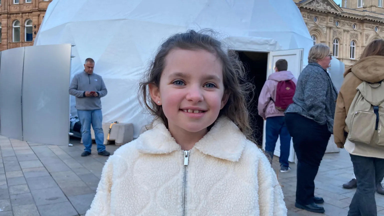 Chloe has long brown hair and is wearing a cream-coloured, fuzzy zip‑up jacket. She stands smiling outdoors in front of a large white dome structure. Several adults are walking or standing in the background on a paved square, with historic buildings visible behind the dome.