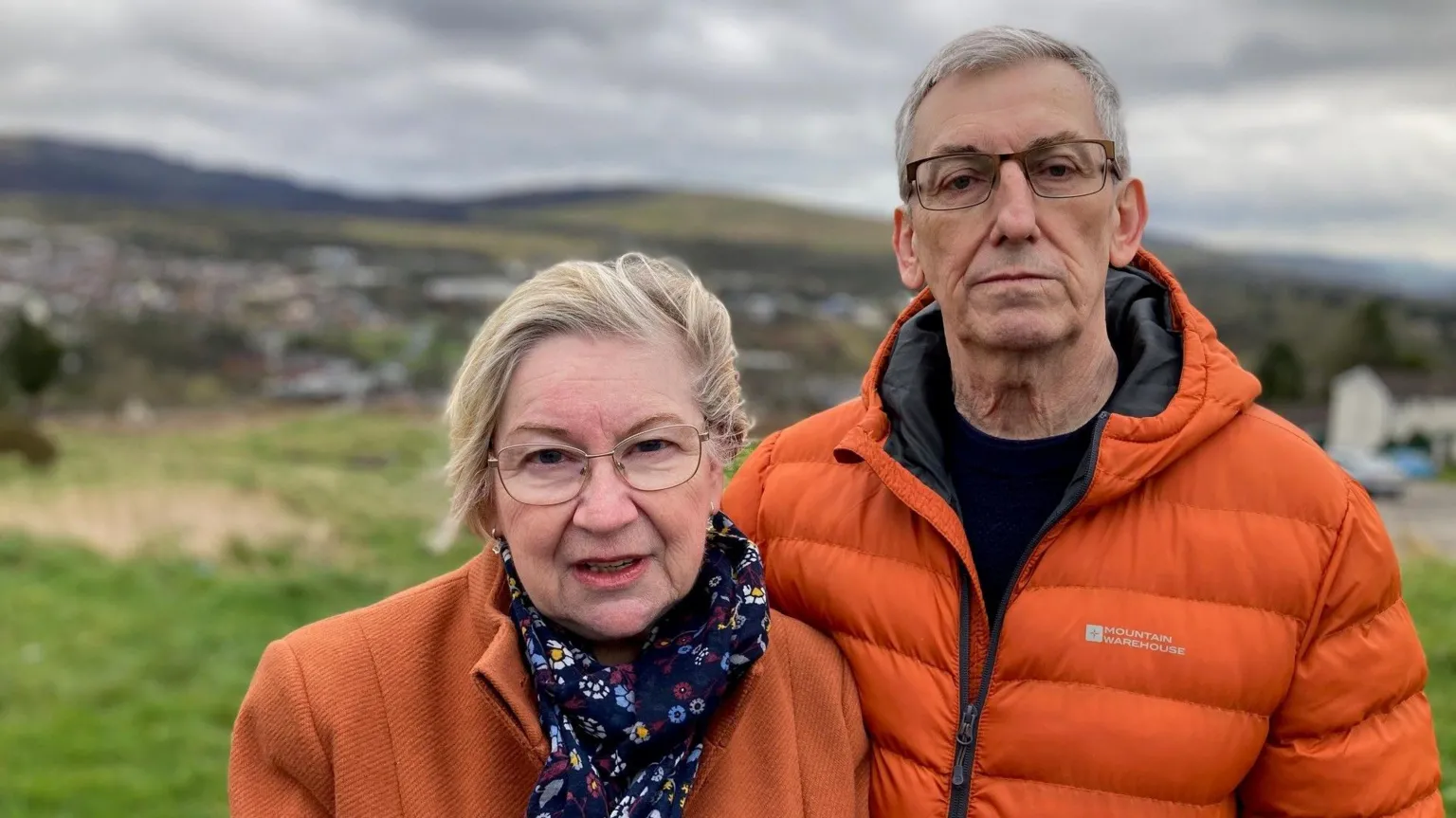 A couple stood outside on a green hill. The background of houses can be seen behind them but is blurred. There is a woman on the left in an orange coat and blue scarf with blonde hair. The man is also in an orange bomber jacket. They both have glasses on and look at the camera.