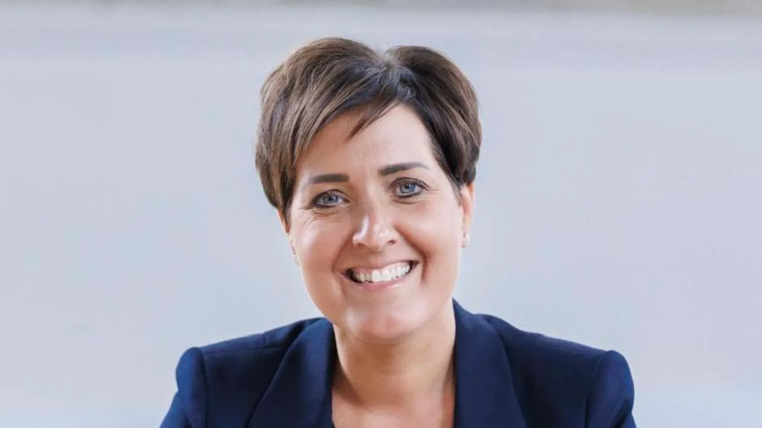 A head shot of a woman with short brown hair in a navy blazer smiling to the camera. The background is white.