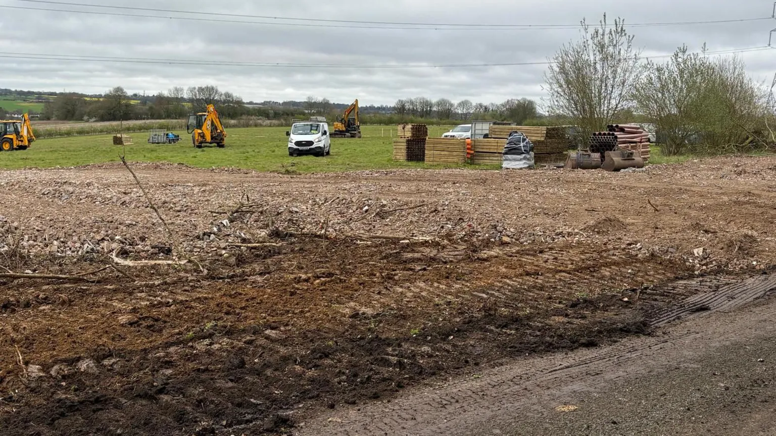 A field, which has been partially turned over to soil, has a series of yellow diggers on. To one side large brown pipes are stacked up alongside wooden beams. Pylon wires run overhead.