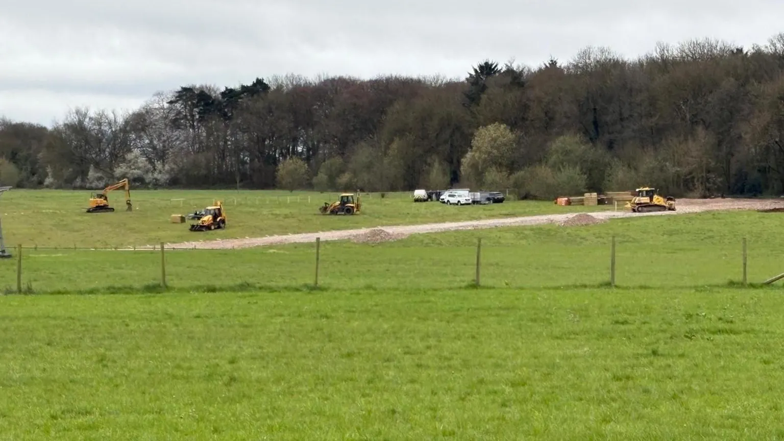 A green field with a barbed wire fence through the middle. At the back part of the field, several diggers roam around and there are piles of gravel stacked up.