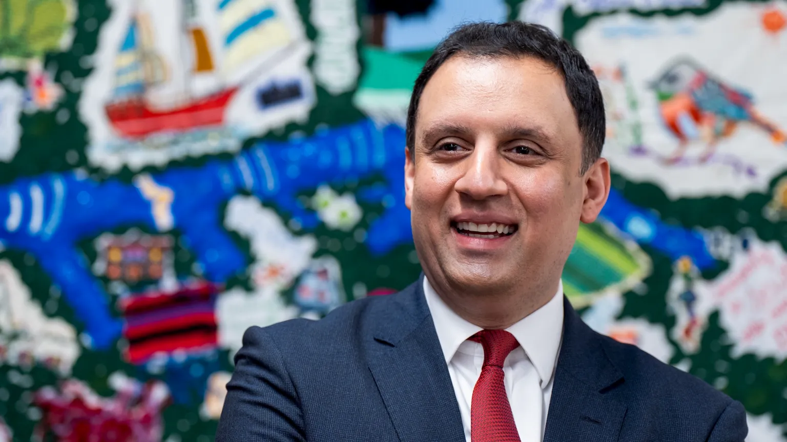  Anas Sarwar in blue suit with white shirt and red tie, stands in front of a colourful backdrop. He is smiling.