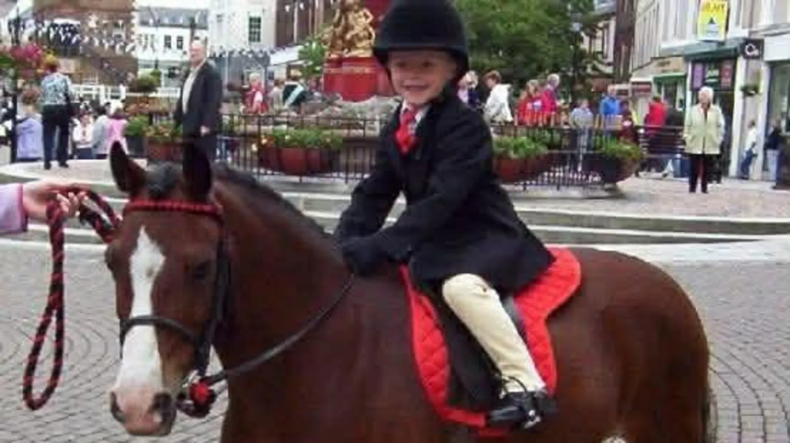 Findlater family Three year old Ross Findlater dressed in a dark riding jacket, hat and jodphurs, sitting on a small brown pony with a white blaze on its face, being held on a leadrope on a cobbled street in Dumfries