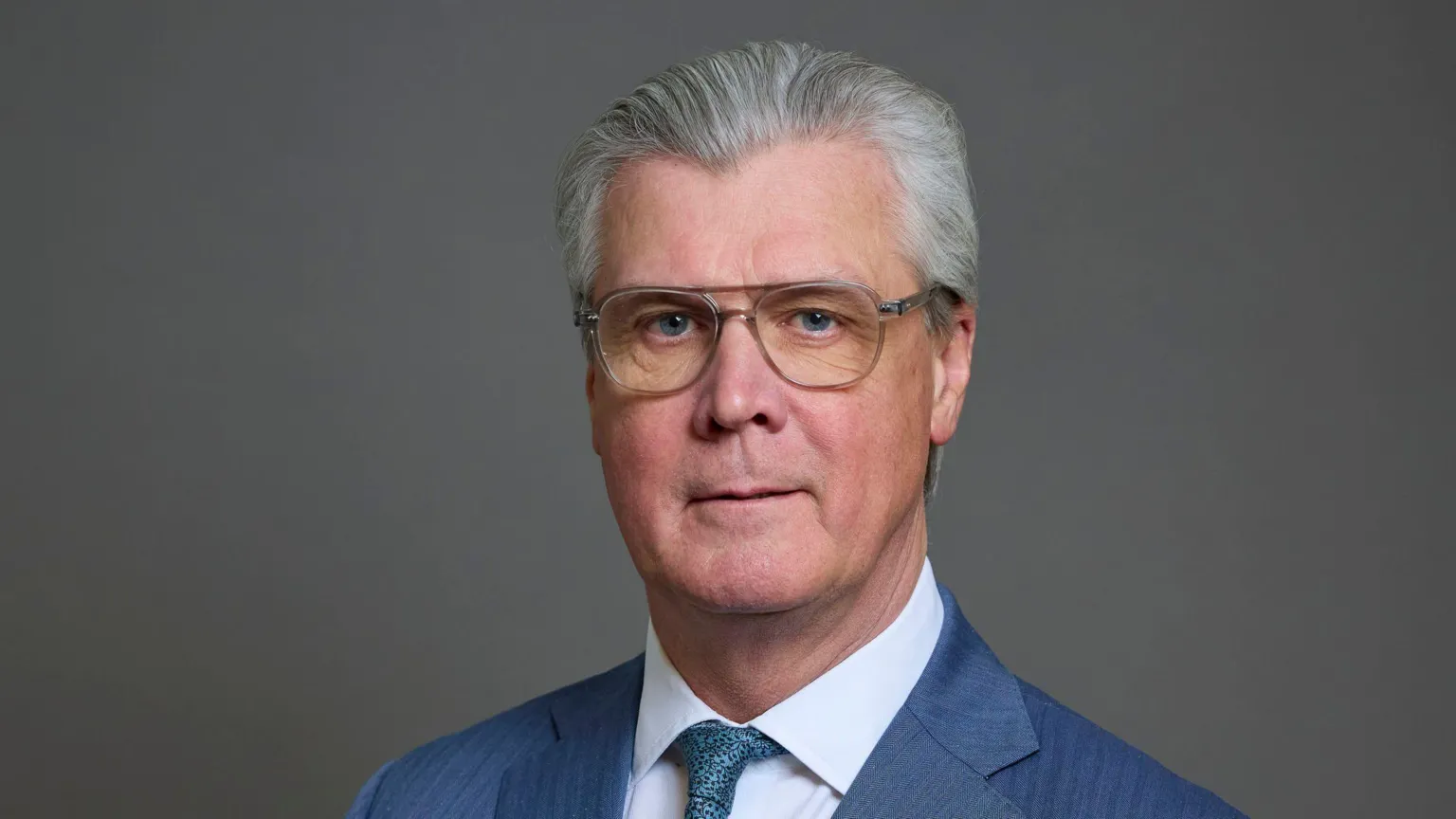  Malcolm Offord has grey hair and glasses. He has a neutral expression and is posing for a portrait style picture against a grey background 