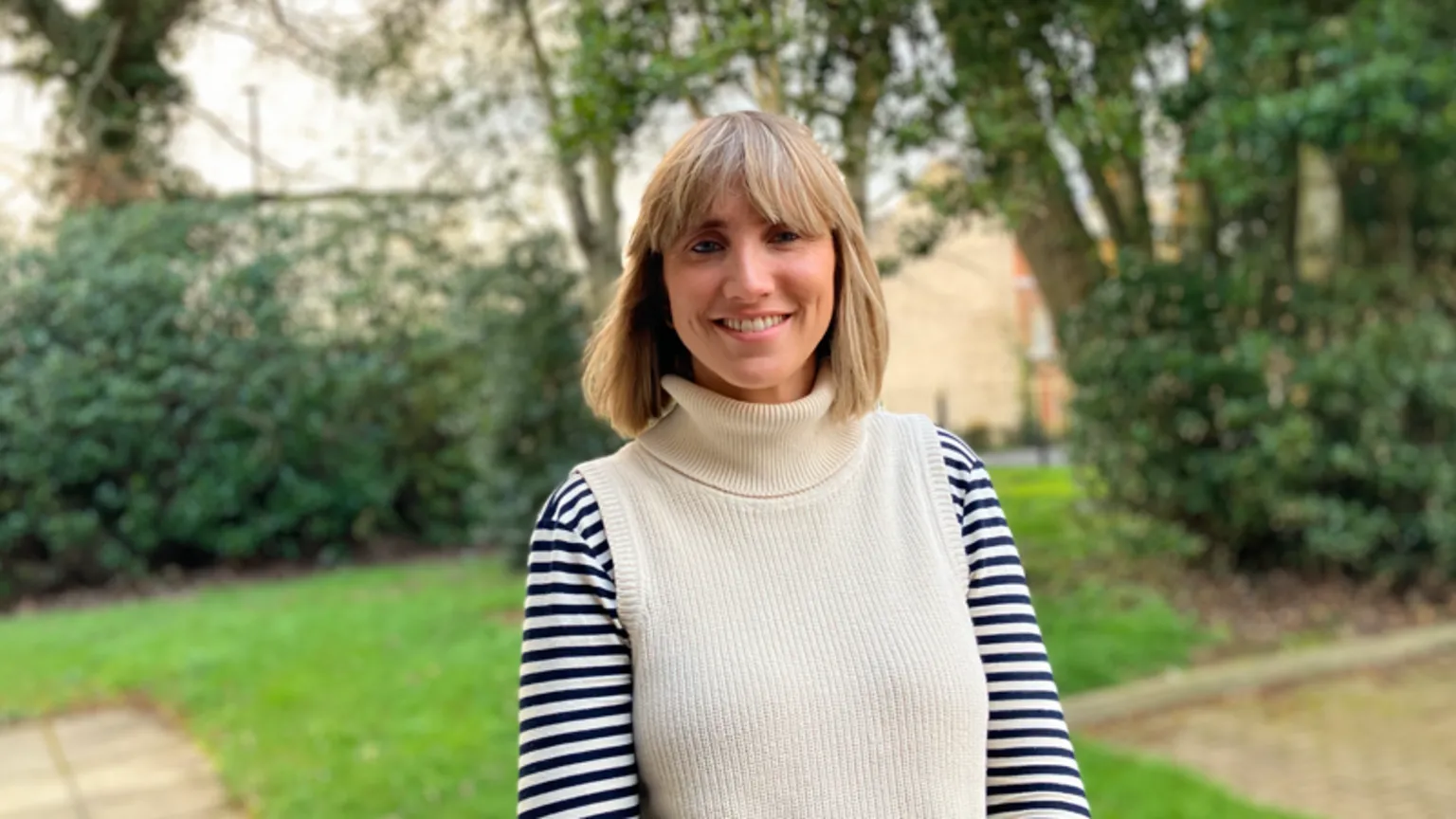 Shows a woman with blonde hair smiling. She is standing in a garden with pavement, grass and trees in the background. She is wearing a roll-neck jumper, which is cream in the middle, with cream and blue striped sleeves.