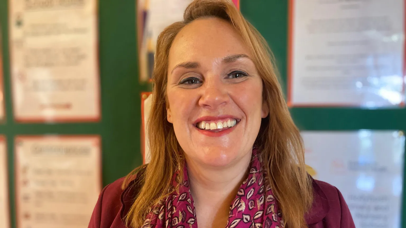 Shows a woman with strawberry blonde hair and smiling in a classroom. She is wearing a burgundy cardigan and a Paisley-design burgundy scarf.
