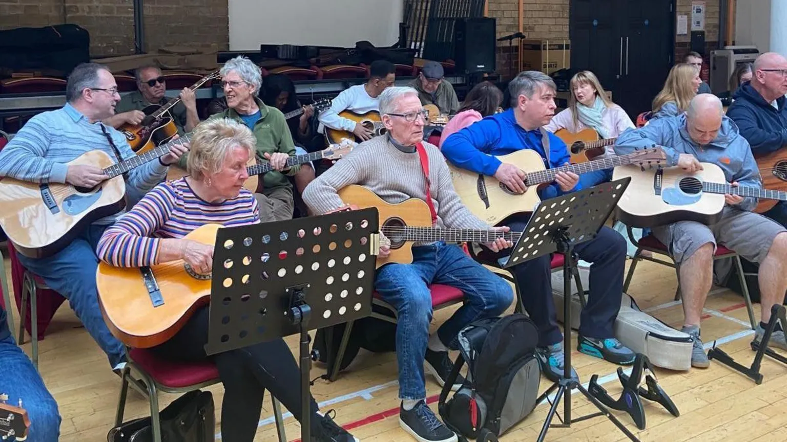 An ensemble of guitarists of all ages sit in two lines at a club with music stands in front of some of them.