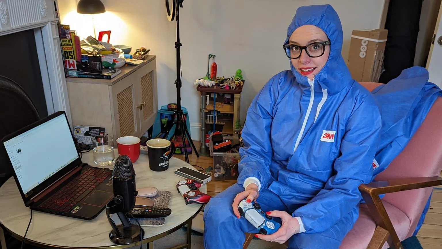 Helen Thorn The comedian Ellie Gibson, who is sitting down inside her flat and wearing glasses and is dressed up from head to toe in a light blue cleaning suit. She is holding a PlayStation controller and next to her on a desk is a small round table with a black microphone and open laptop as well as mugs of tea.