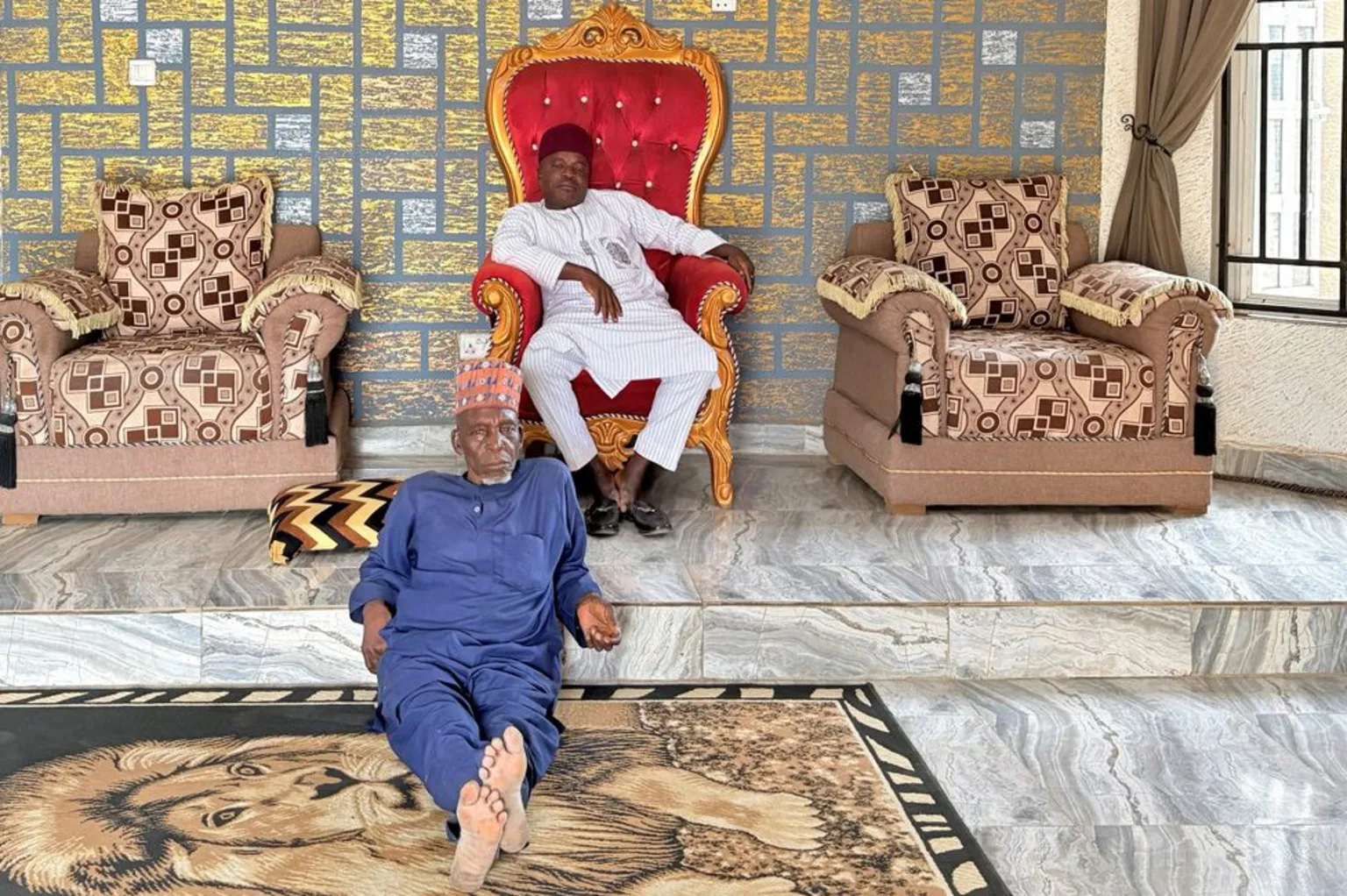Adaobi Tricia Nwaubani Chief Pada sitting on his red and gold throne in his palace in Mariana-Kubwa. Sitting on the floor is John Ngbako on a rug with a lion design on it.