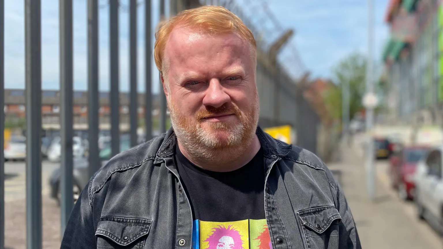 Brian Smyth has ginger hair and beard and is looking at the camera, he is wearing a black denim jacket and T-shirt and is standing next to a metal fence. Behind him out of focus is a street scene in Belfast with parked cars.