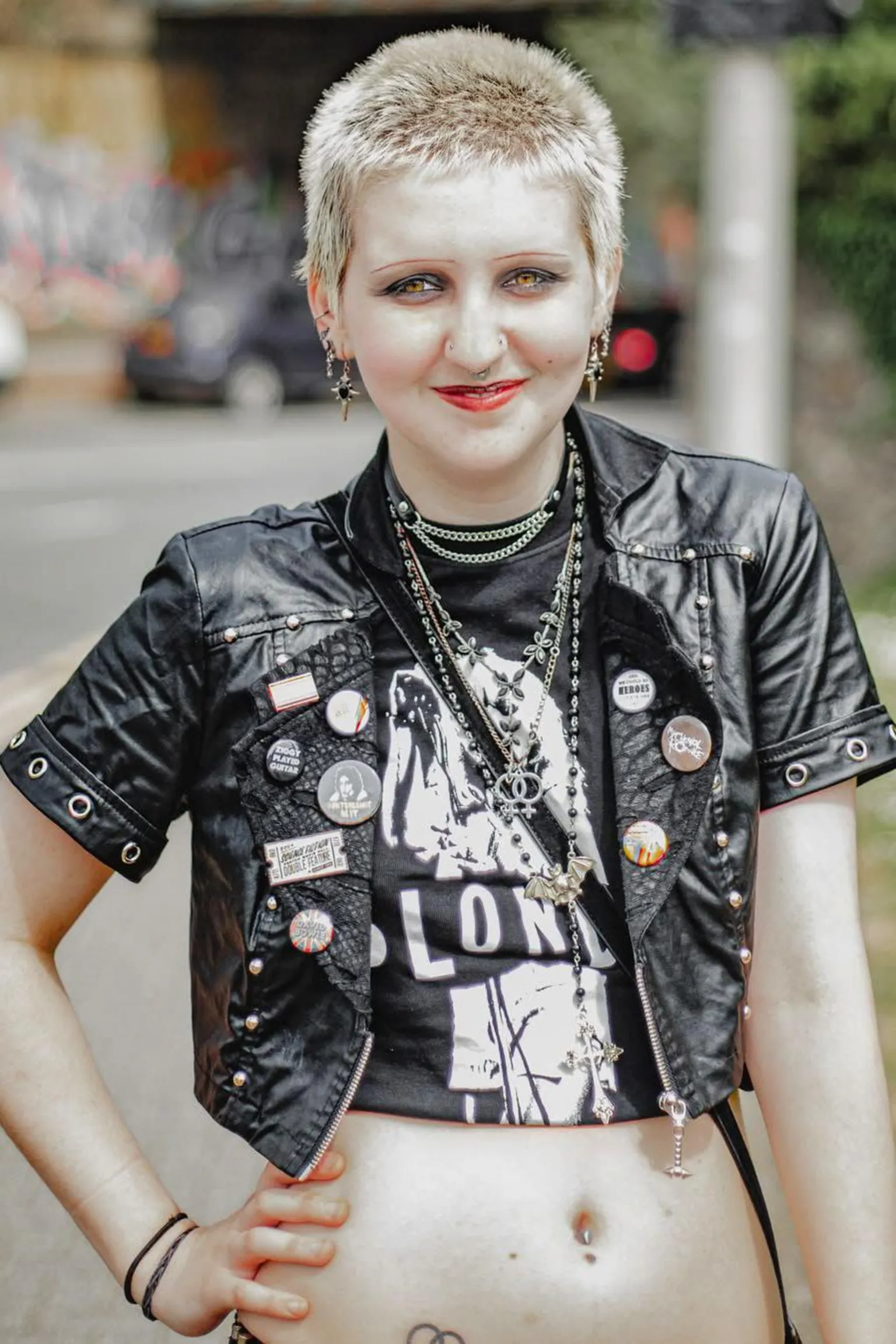 Tom Wilkins Deena stands on a pavement in the street. She has blonde short hair in a buzz cut, wears black eyeliner and red lipstick. She holds her left hand to her hip and smiles at the camera. Her belly is on show and she wears a black cropped t-shirt and black cropped leather jacket. She has lots of silver necklaces layered on top of her top. 