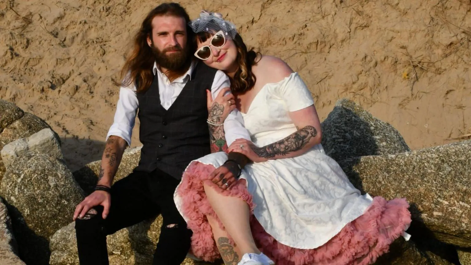 Tom Wilkins Tom Wilkins sits on some rocks in the sand on a beach next to his wife. He wears a waistcoat and trousers and a white shirt and looks at the camera. He has long brown hair and a thick beard. His wife leans into his shoulder and holds his one 