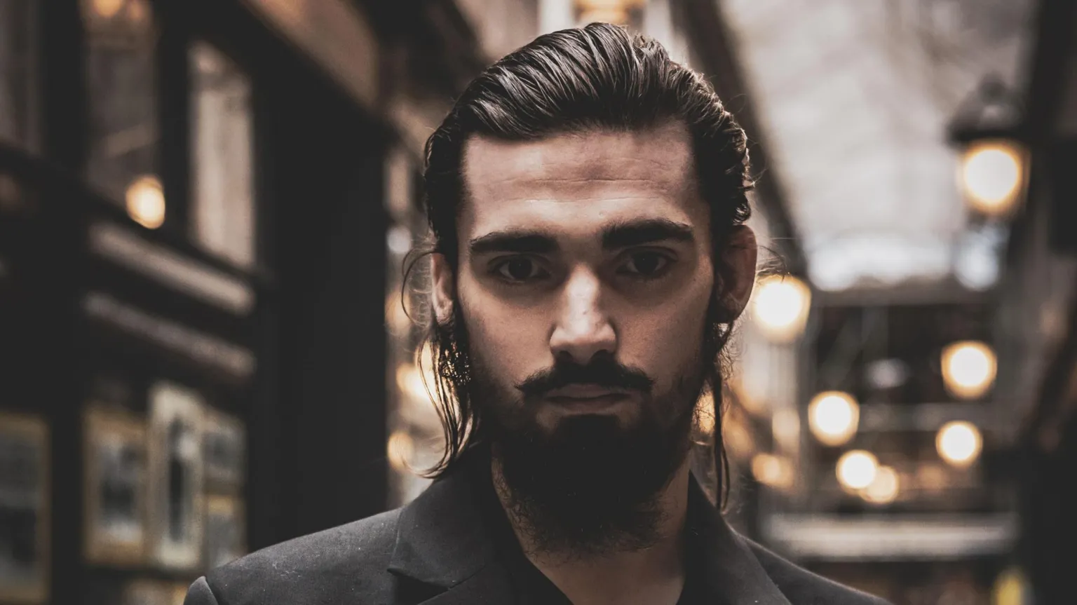 Tom Wilkins Theo, one of Tom's subjects, is pictured inside Cardiff's arcades. He has brown eyes and a brown beard, with long hair pulled back. Lights can be seen in the background but it's blurred. 