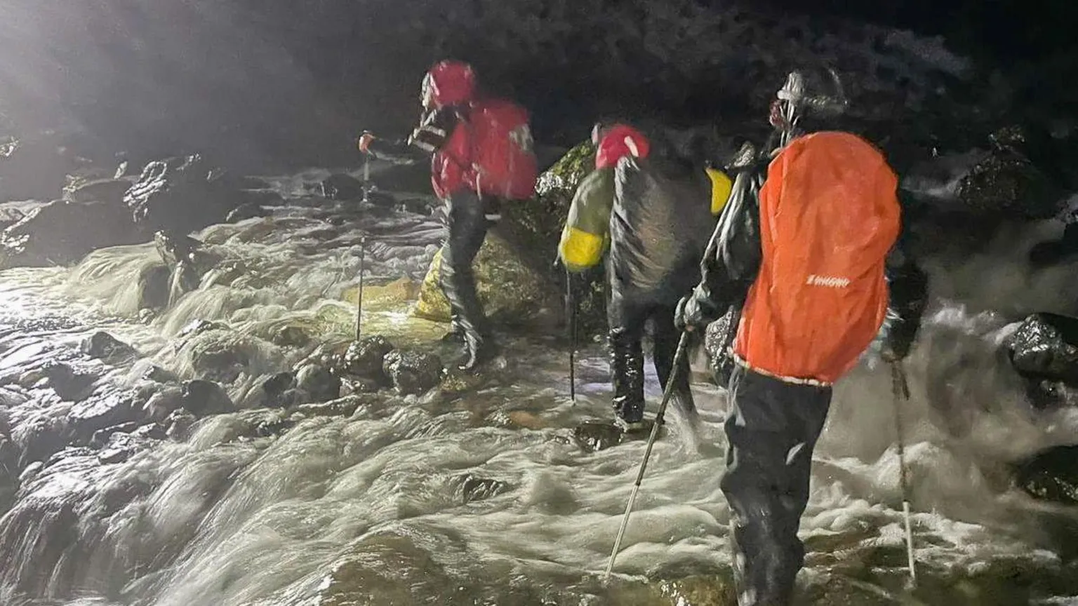WASDALE MOUNTAIN RESCUE TEAM Mountain rescue volunteers make their way over a waterfall which is in spate due to the harsh weather conditions. The volunteers look soaked through and their waterproofs are shining with how wet they are. The water is lit up by headtorches as it is at night.