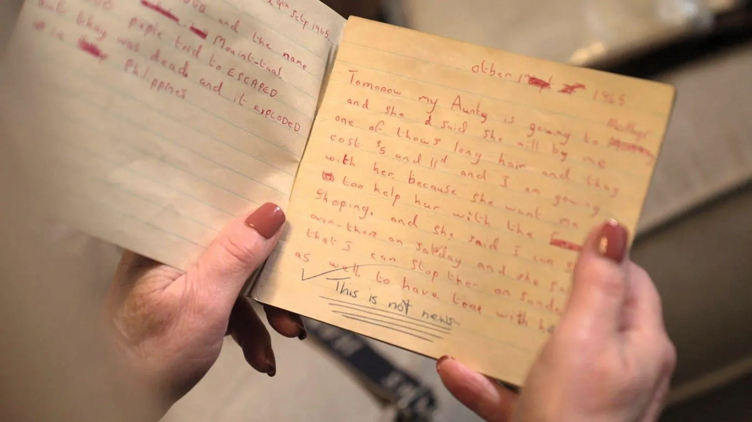 A close up of a school exercise book from the 1960s, the pages are yellowed. One page is filled with text handwritten in red ink, which details a trip to the shops. At the bottom of the page, underlined, a teacher has written