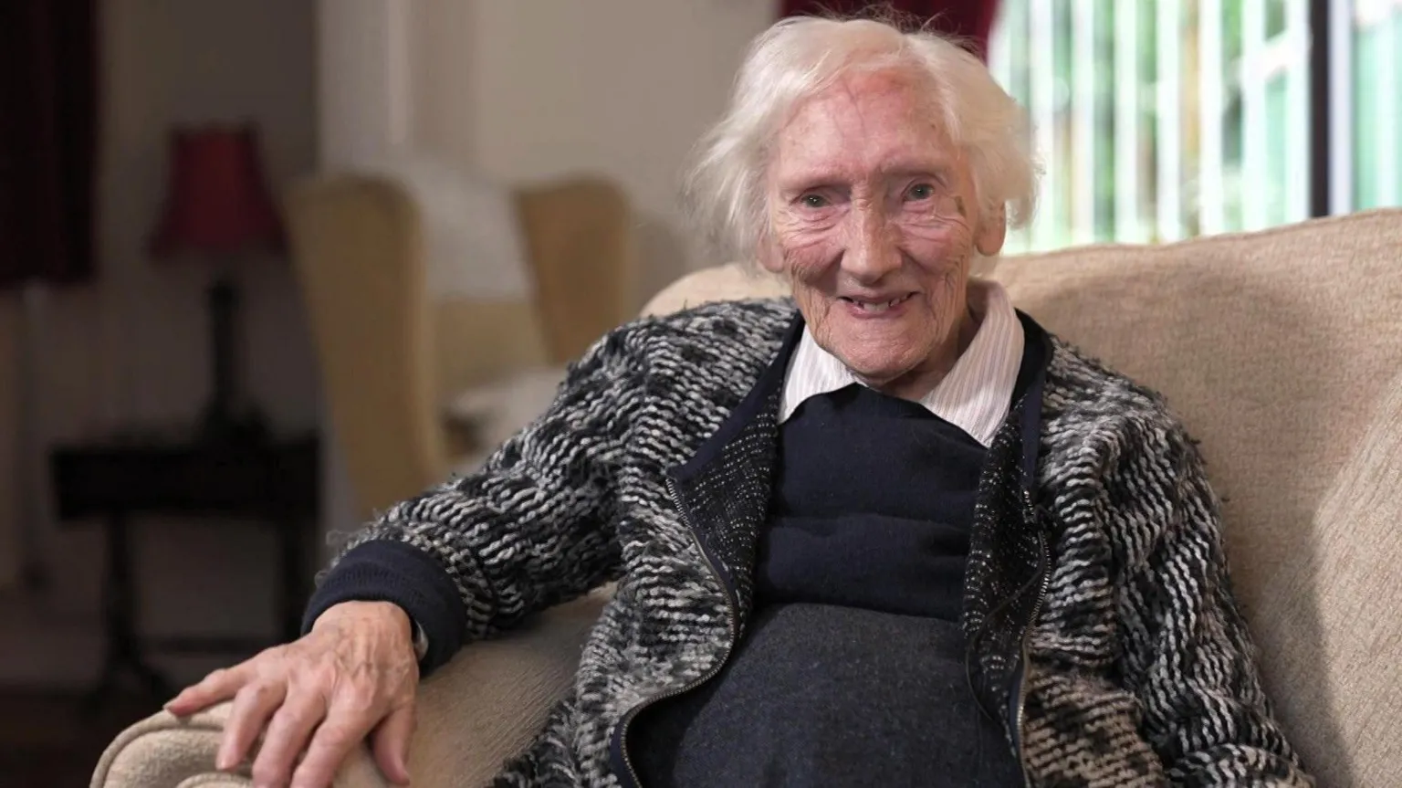 A woman with short white hair looks at the camera. She has a black and white knitted cardigan and has a black smock over a white collared shirt - the collars can be seen. She is sat on a cream sofa. She is in focus while the background is blurred.
