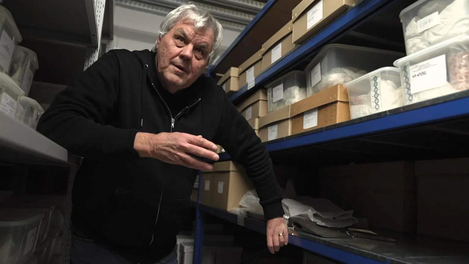 A man with grey hair and a black hoody looks towards the camera. He is surrounded by boxes on shelves.