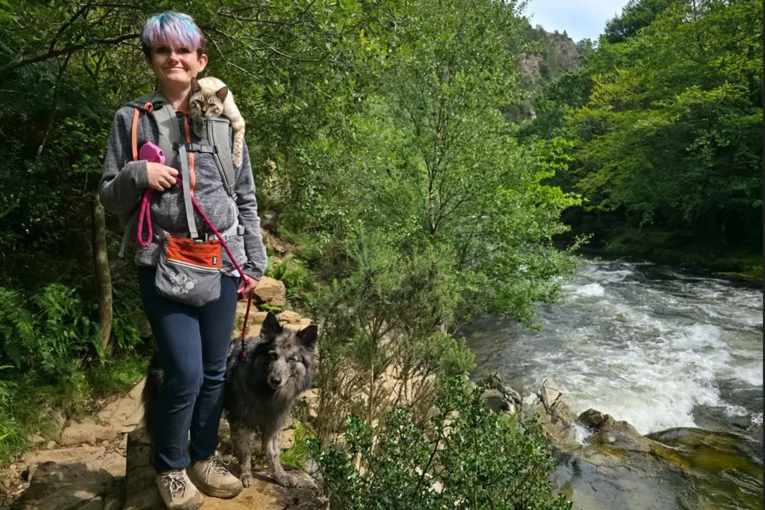 Candice Stapleton A woman with short purple and blue dyed hair stands on a river bank. There is a cat lying on her shoulder and a dog stood at her side.
