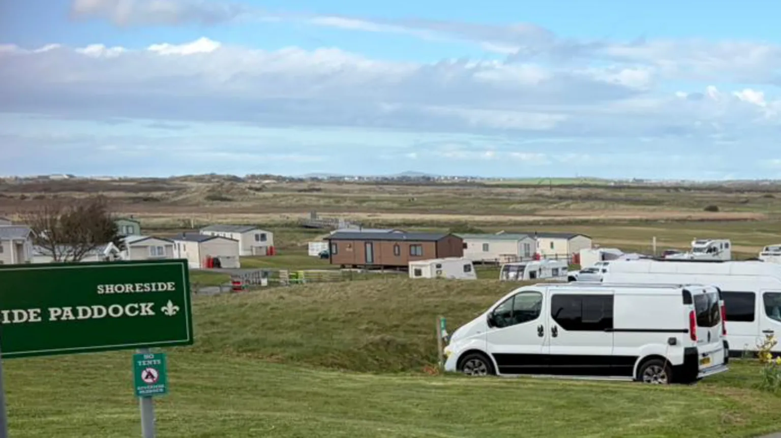 Grassy banks with parked campervans, motorhomes, and static caravans arranged in neat rows. A green sign on the left reads “Shoreside Paddock,” marking part of the site. Wooden fencing and a metal gate line the right side of the road, with a tall utility pole nearby. In the distance, flat fields stretch toward the horizon under a bright blue sky with scattered clouds, giving the scene a spacious, coastal feel.