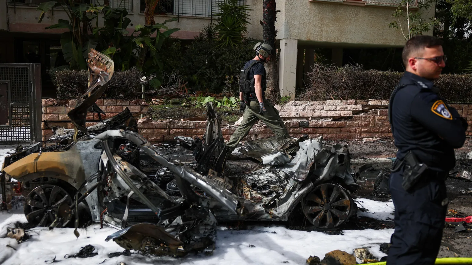 Uniformed member of Israeli security services stands in the right of the picture looking right. Beside him is the wreckage of a vehicle and behind it another man is walking.
