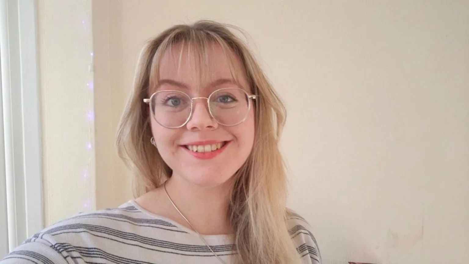 Catherine Thoyts A female, smiling, wearing glasses and a striped white and grey top, with a silver necklace. She is standing in front of a cream-coloured wall.