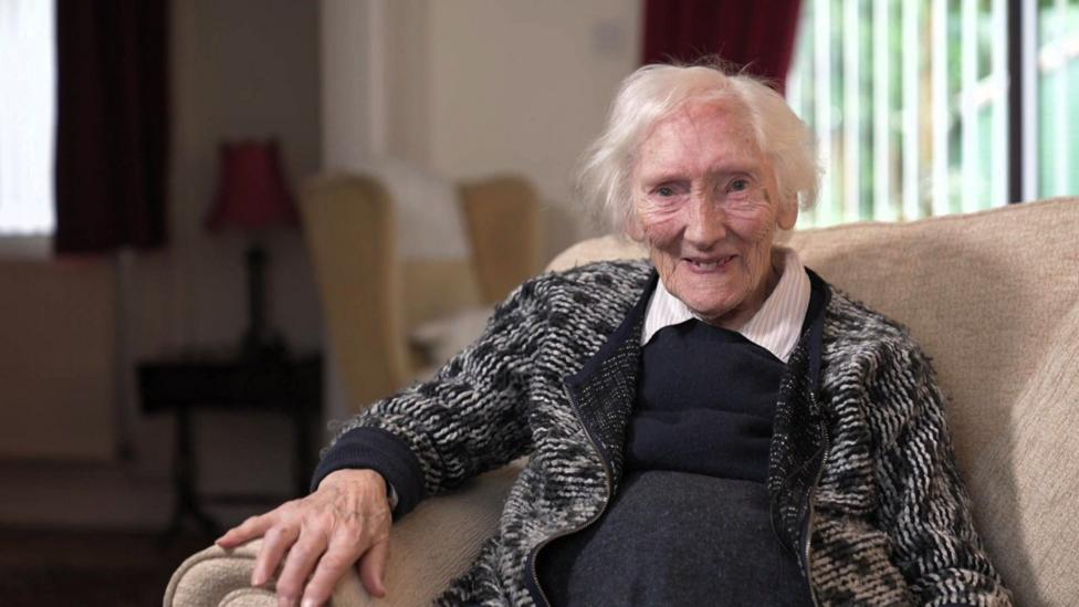 Menyw mewn oed gyda gwallt llwyd a gwyn, yn gwenu tuag at y camera. Mae hi'n eistedd ar sofa lliw golau mewn lolfa taclus.