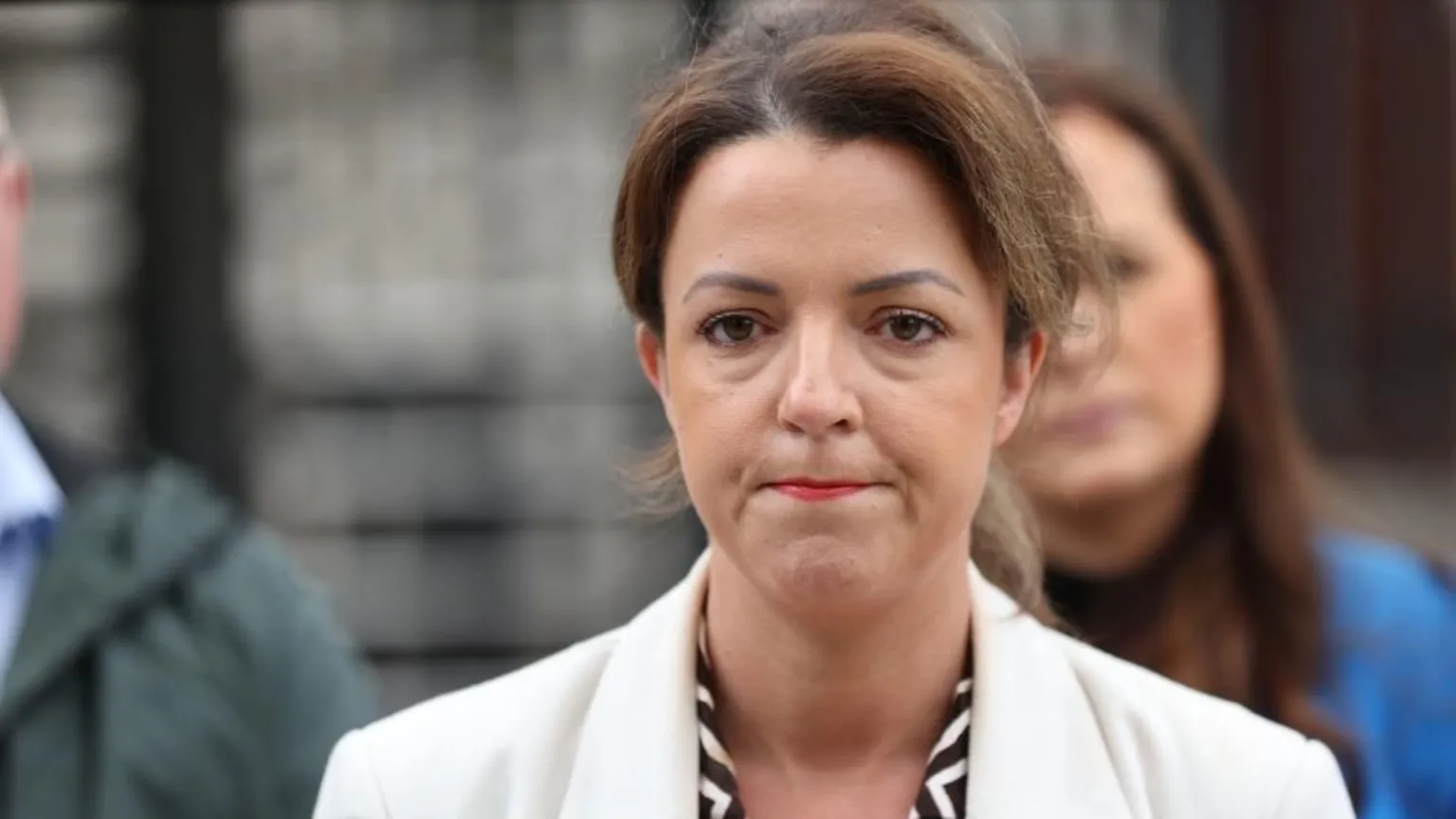 A woman with brown hair looks into the camera. She is wearing a white jacket and black and white patterned shirt.