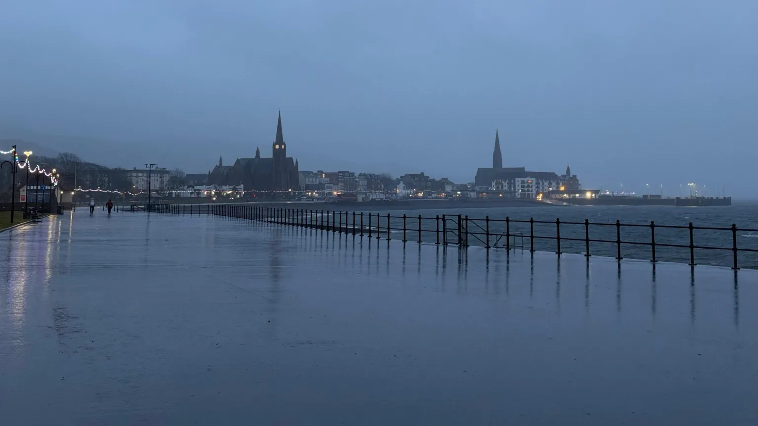 BBC Weather Watchers/Lawrence A dark view of a promenade by a beach. It is covered in water and the lights of a town can be seen in the distance