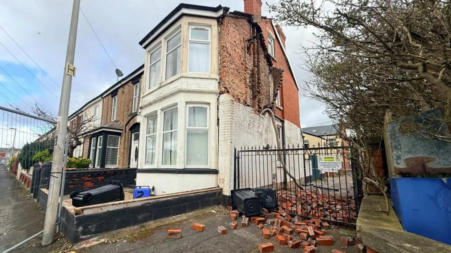 A pile of red bricks are scattered across a gated alleyway and the pavement, with a hole left in the side of the wall on the first floor wall of the house. The area is fenced off.