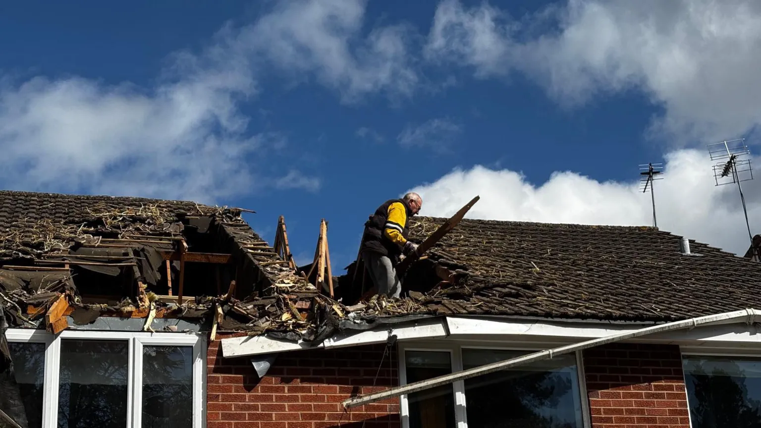 A man in a yellow top stands in a large hole in the roof of two houses