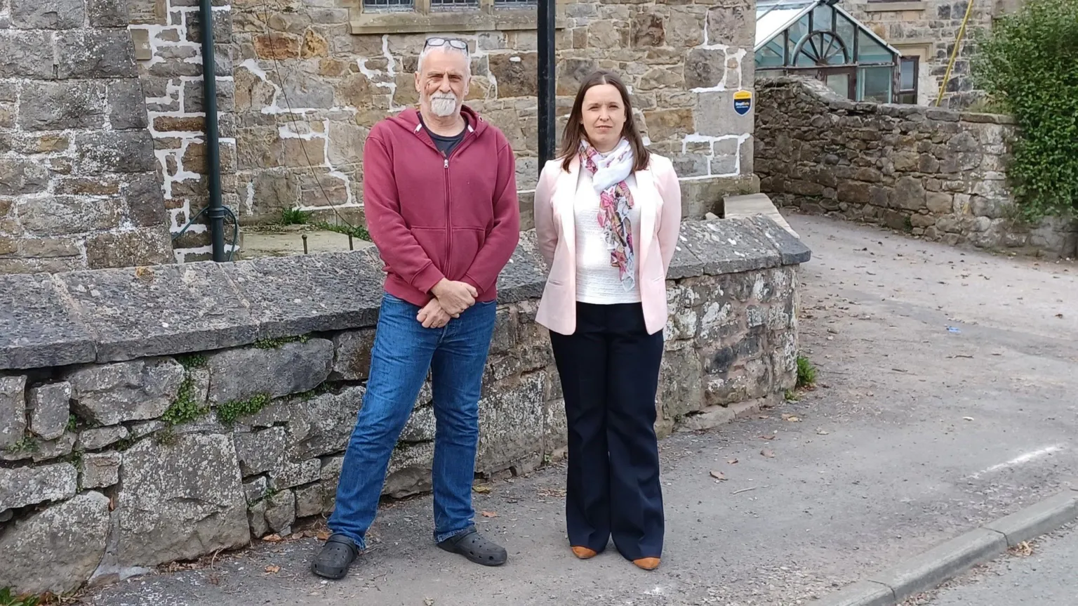 A man and a woman stood on the pavement outside a stone house.