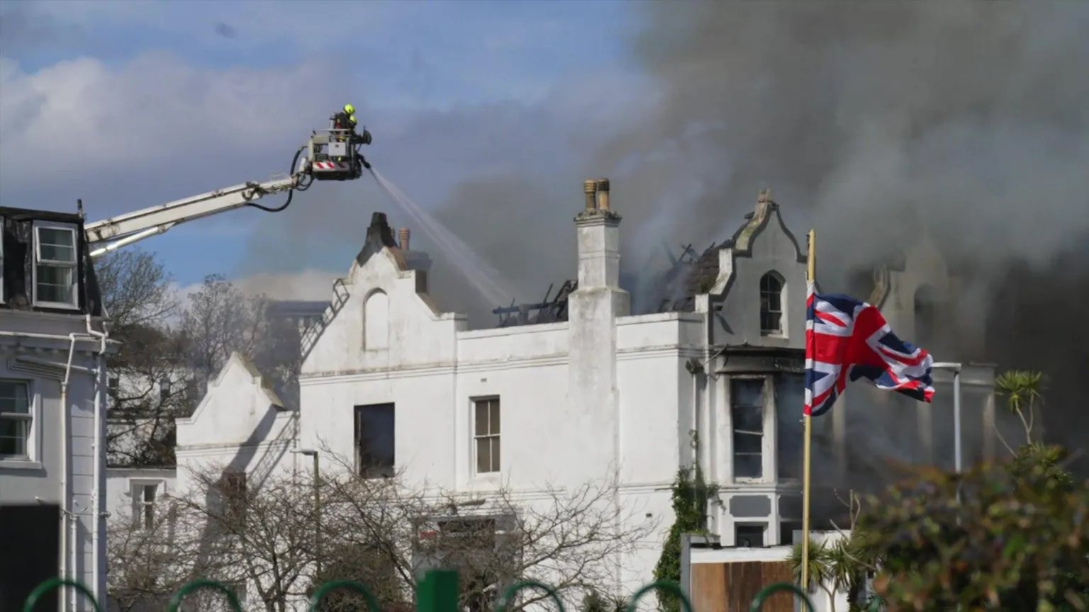 Black smoke coming from the derelict hotel in Torquay. The smoke is black and is filling the sky. A firefighter is on an ariel ladder to the right fighting the fire.