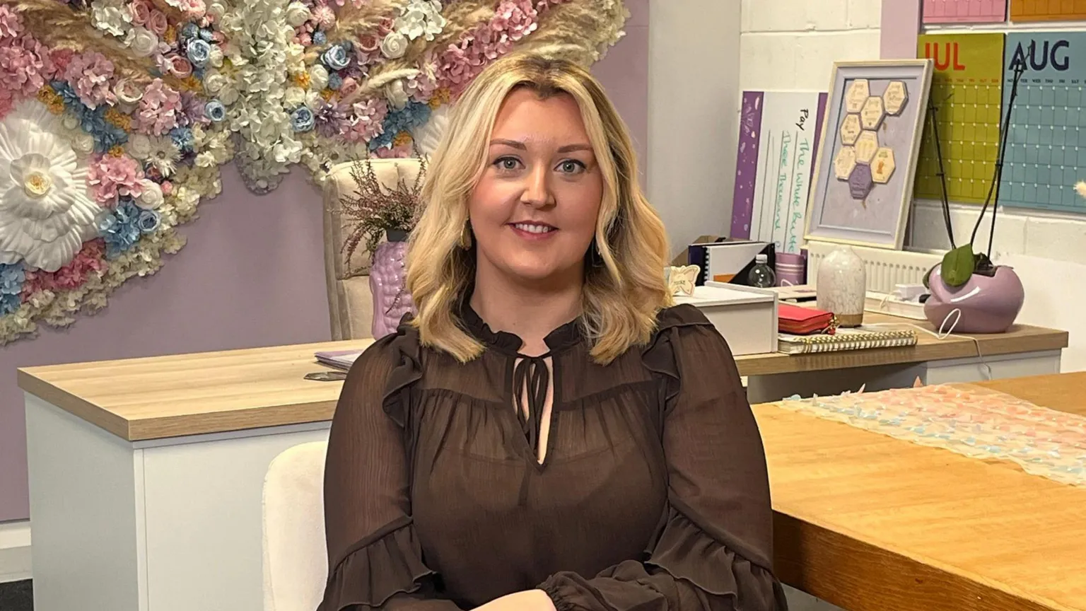 Rebecca is sitting on a chair. She is wearing a black top and has medium blonde hair. To her right is a table. There is a desk behind her. The walls are decorated in colour. 