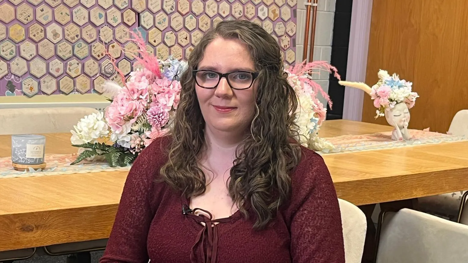 Victoria is sitting. Behind her is a wooden table. She is wearing a burgundy top and has long brown hair. She is wearing black glasses. The wall of tiles is behind her. There are flowers on the table. 