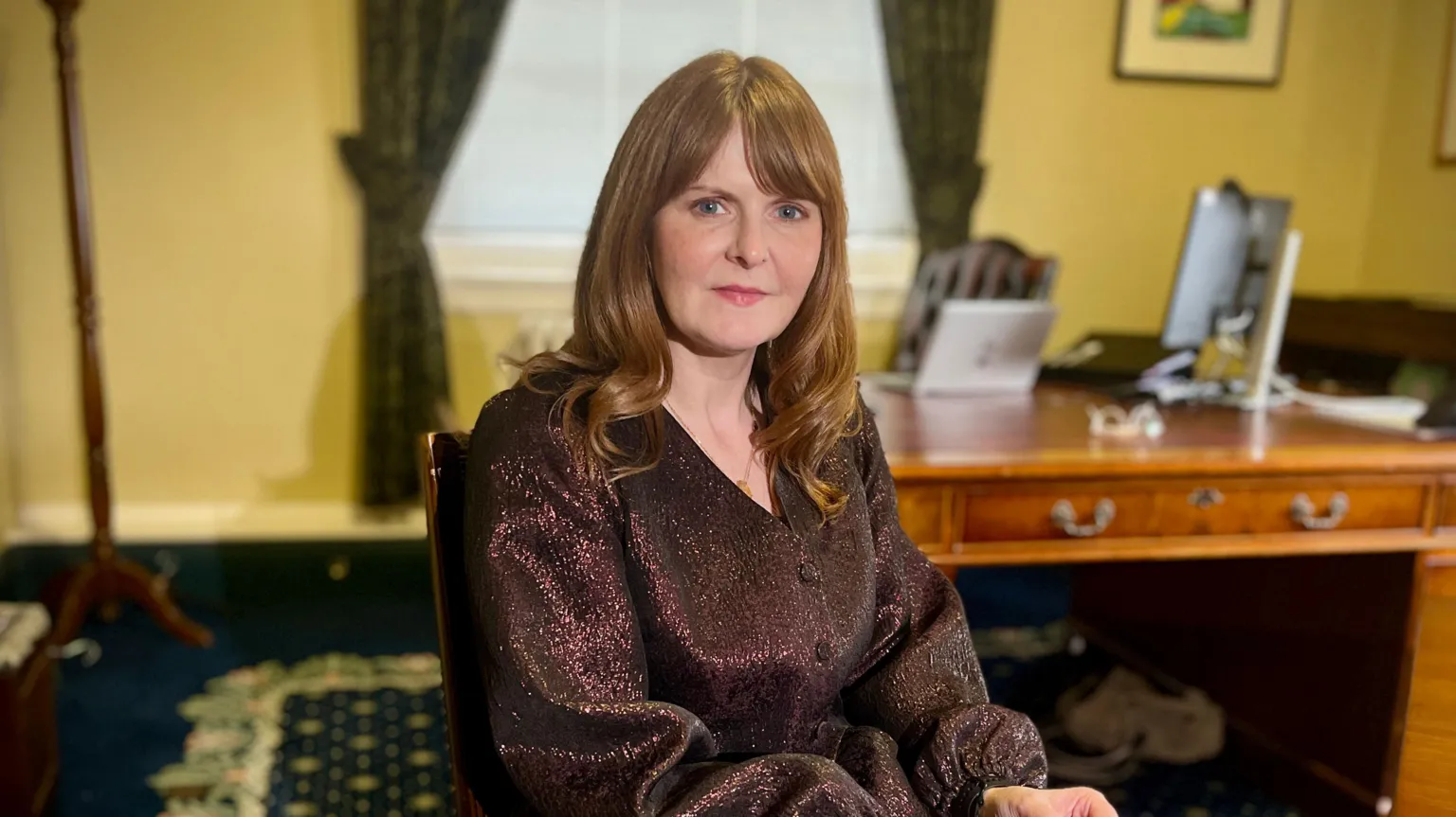 Caoimhe is sitting. Behind her is a brown desk and a yellow coloured wall. She is wearing a sparkled black shirt. She has medium brown hair and a chain around her neck. The carpet in the room is navy. 
