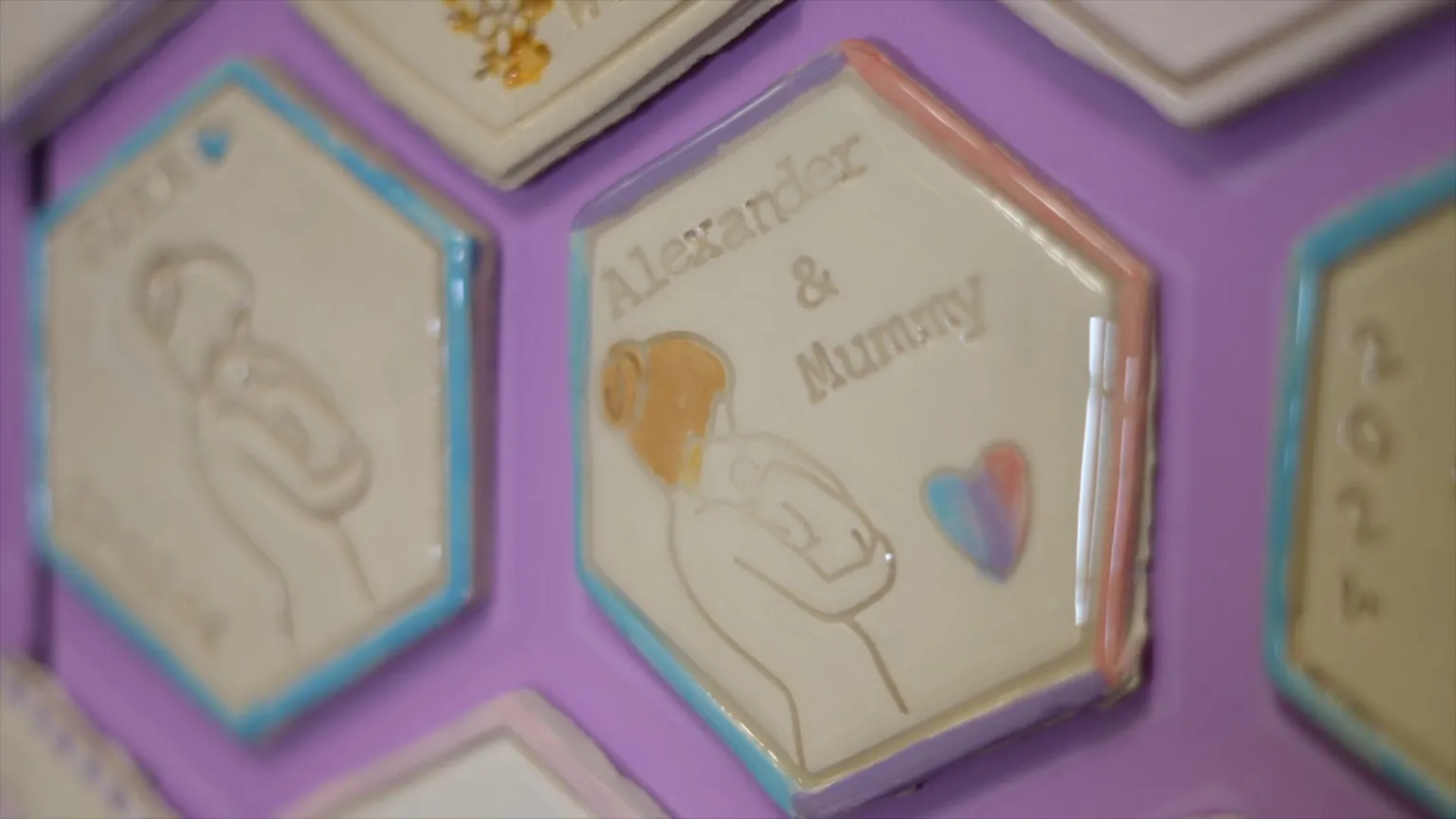 A single ceramic tile is focused, the rest are blurry. The tile says Alexander & Mummy on it. The wall is purple. 
