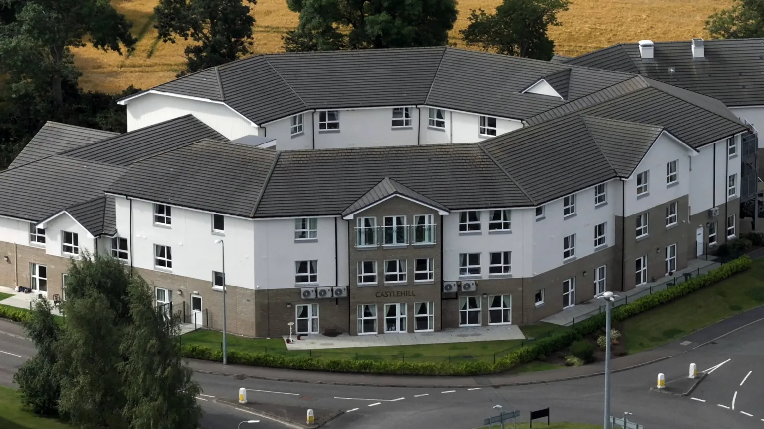 An exterior shot of a large modern care home. It is a grey and white building with three floors and rows of windows in its walls. There is a road with a roundabout near the main entrance.