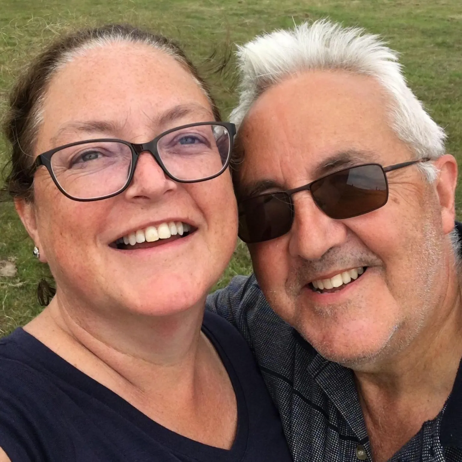 A smiling man and woman taking a close-up selfie outdoors with grassy cliffs in the background.