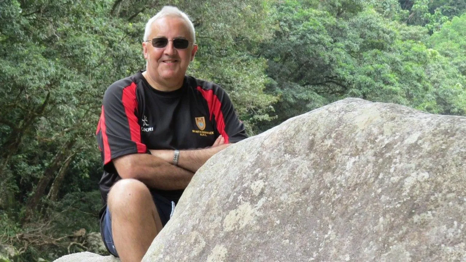 A man sitting outdoors on large rocks, smiling with arms folded, wearing sunglasses and a black and red sports shirt, surrounded by green trees.
