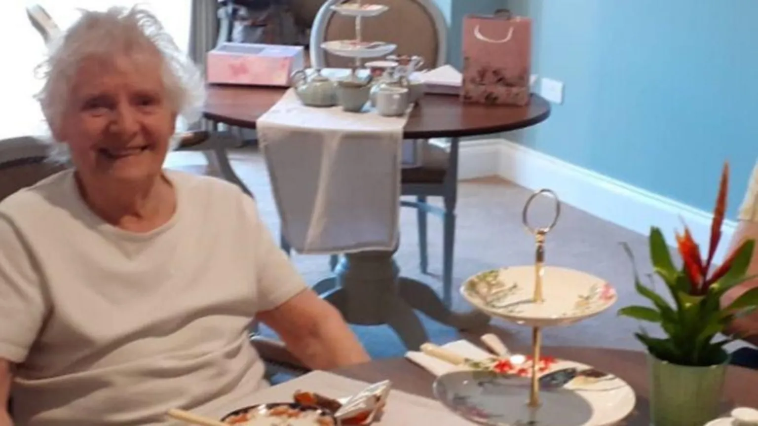 A smiling woman sitting at a table set for afternoon tea, with tiered cake stands, teapots, and a potted plant in a bright, nicely decorated room.