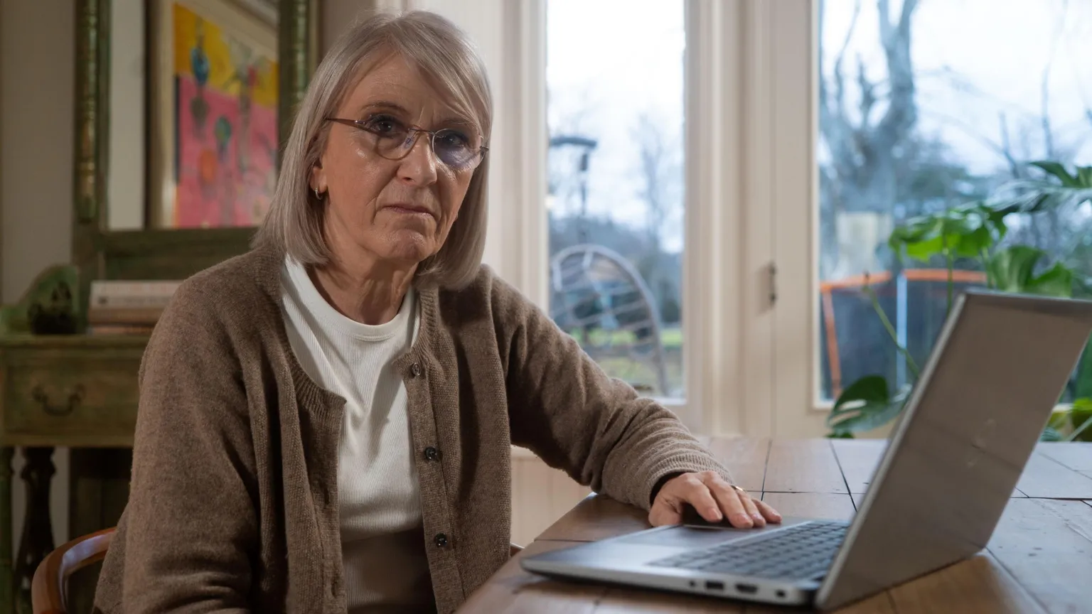A woman sitting at a wooden table with a laptop, looking toward the camera, in a bright room with large windows and artwork in the background. 