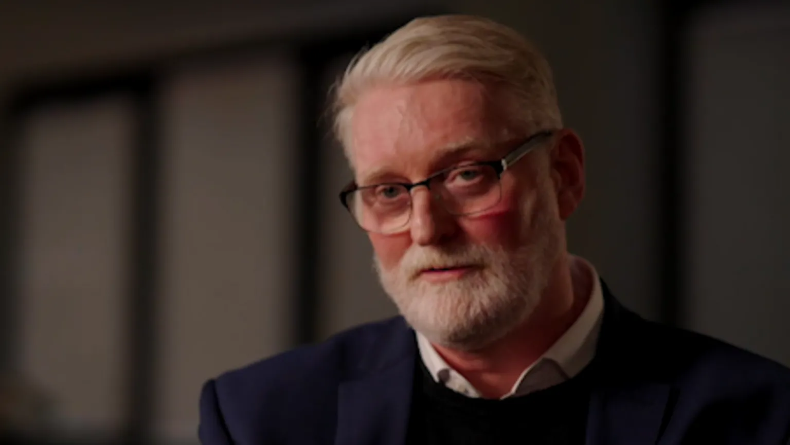 Jim Pearson, a man with grey hair and a grey beard, looking at the camera. He is wearing glasses, a white shirt, a black jumper and a dark blue jacket.