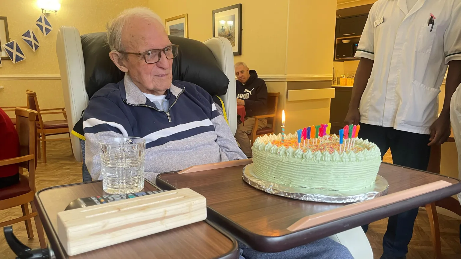 Elderly man sitting in a care‑home chair with a birthday cake and lit candles on the tray in front of him, as staff and residents look on in the background.