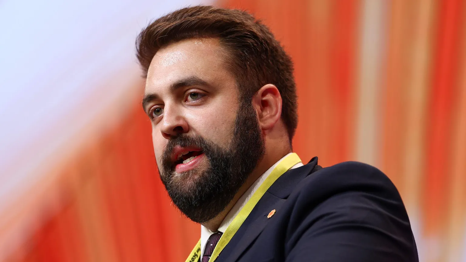 Plaid economy spokespersons Luke Fletcher wearing a suit. It is a head and shoulders shot and he is stood in front of an orange background.